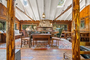 Interior d'una cabana de fusta amb una llar de pedra, mobles de fusta i grans finestres.