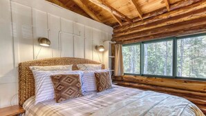 A log cabin bedroom with a bed, two wall-mounted lights, and a window with a forest view.