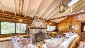 A log cabin interior with a stone fireplace, wooden furniture, and large windows.