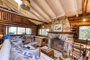A log cabin interior with a stone fireplace, wooden furniture, and large windows.