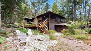A cabin with a deck, surrounded by trees, and two Adirondack chairs on a stone pathway.