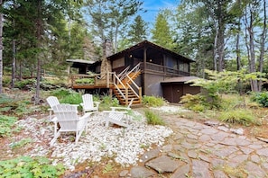 A cabin with a deck, surrounded by trees, and two Adirondack chairs on a stone pathway.