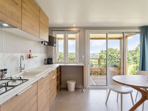 A modern kitchen with wooden cabinets, a gas stove, and a sink. There is a small trash bin and a window overlooking a balcony with a view of greenery. - Camping Papillon Bouillon - Vakantiehuis Premium by Interhome (Bouillon)