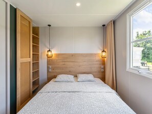 A bedroom with a wooden headboard, two bedside lamps, a bed with patterned bedding, a wardrobe with a woven door, and a window with a view of greenery. - Camping Papillon Bouillon - Vakantiehuis Premium by Interhome (Bouillon)