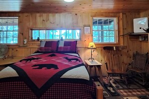 A wooden cabin room with a bed, two windows, a lamp, a chair, and a fan.