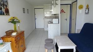 Una sala de estar con un sofá azul, una mesa blanca y una cocina al fondo.