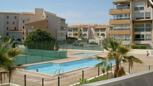 Zona de piscina con terraza amurallada, palmeras y edificios de varios pisos al fondo.
