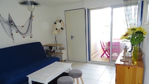 Una sala de estar con un sofá azul, una mesa de centro blanca, dos taburetes grises, un mueble de madera y vistas a un área exterior con una silla rosa a través de una puerta de cristal.