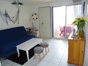 Ein Wohnzimmer mit einem blauen Sofa, einem weißen Couchtisch, zwei grauen Hockern, einer Holzkommode und Blick durch eine Glastür auf einen Außenbereich mit einem pinken Stuhl.