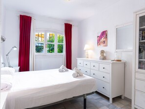 Une chambre d’hôtel avec un lit, une commode, une lampe, un tableau et une fenêtre avec des rideaux.