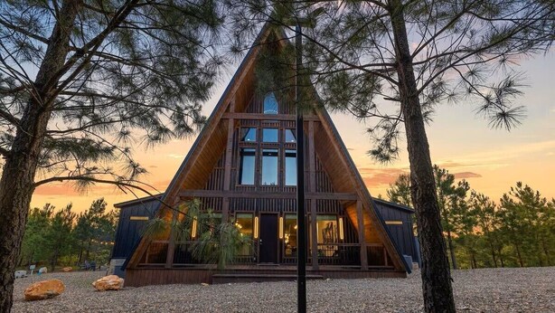 Uma cabana de madeira triangular com uma grande janela na frente e uma porta, rodeada por árvores e um caminho de acesso de cascalho.