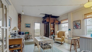 A cozy living room with a dining table, a yellow armchair, and a kitchen area with a counter and stools. - Sunrise on the Sheepscot- condo w/ deck overlooking the water - steps to dining & shopping (Wiscasset)