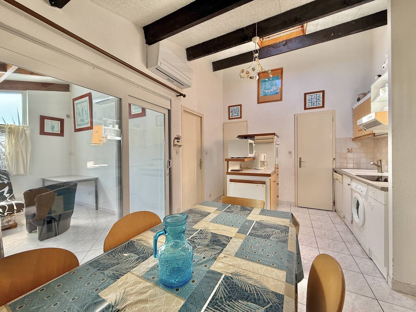 Uma área de refeições com uma mesa, cadeiras, um jarro azul e uma cozinha com eletrodomésticos.