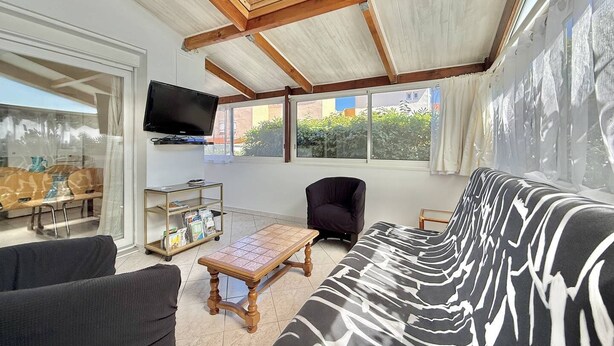 Ein Wohnzimmer mit einer Couch, Sesseln, einem Couchtisch, einem an der Wand montierten Fernseher und großen Fenstern mit Blick auf Grünflächen.
