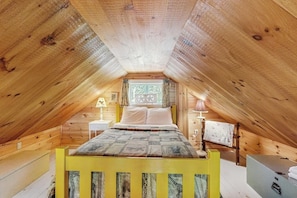 A cozy attic bedroom with a wooden sloped ceiling, a bed with a patterned bedspread, a nightstand, a lamp, and a window with curtains. - Lakefront, cabin-inspired home with firepit, grill, & screened porch, Wifi (Greenville)