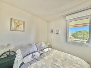 A bedroom with a bed, a nightstand, a lamp, a framed picture, a window with a view, and a thermostat. - CAP D'AGDE - Plage Richelieu / Réf 0154 (CAP D'AGDE)