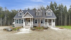 A two-story house with a porch, a small front yard with rocks, and a forest in the background.