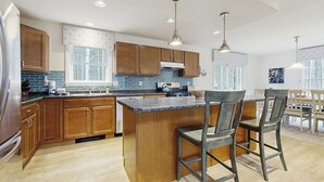 A modern kitchen with wooden cabinets, a central island, bar stools, pendant lighting, and a view of the dining area.