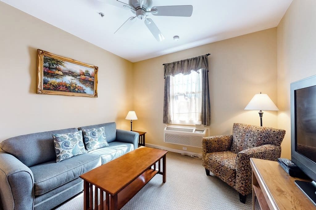 A living room with a sofa, armchair, coffee table, TV, ceiling fan, air conditioning unit, and a painting on the wall.