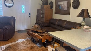Ein Wohnzimmer mit braunem Ledersofa, einem Holztisch, einem Kuhfellteppich, einer Küchenzeile mit Barhocker und einem Deckenventilator.