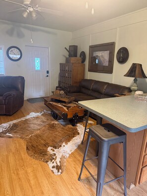 A living room with a brown leather couch, a wooden coffee table, a cowhide rug, a kitchen area with a bar stool, and a ceiling fan. - Blue Sky Bungalow - 1-bedroom bungalow in Homosassa located just off 19! (Homosassa)