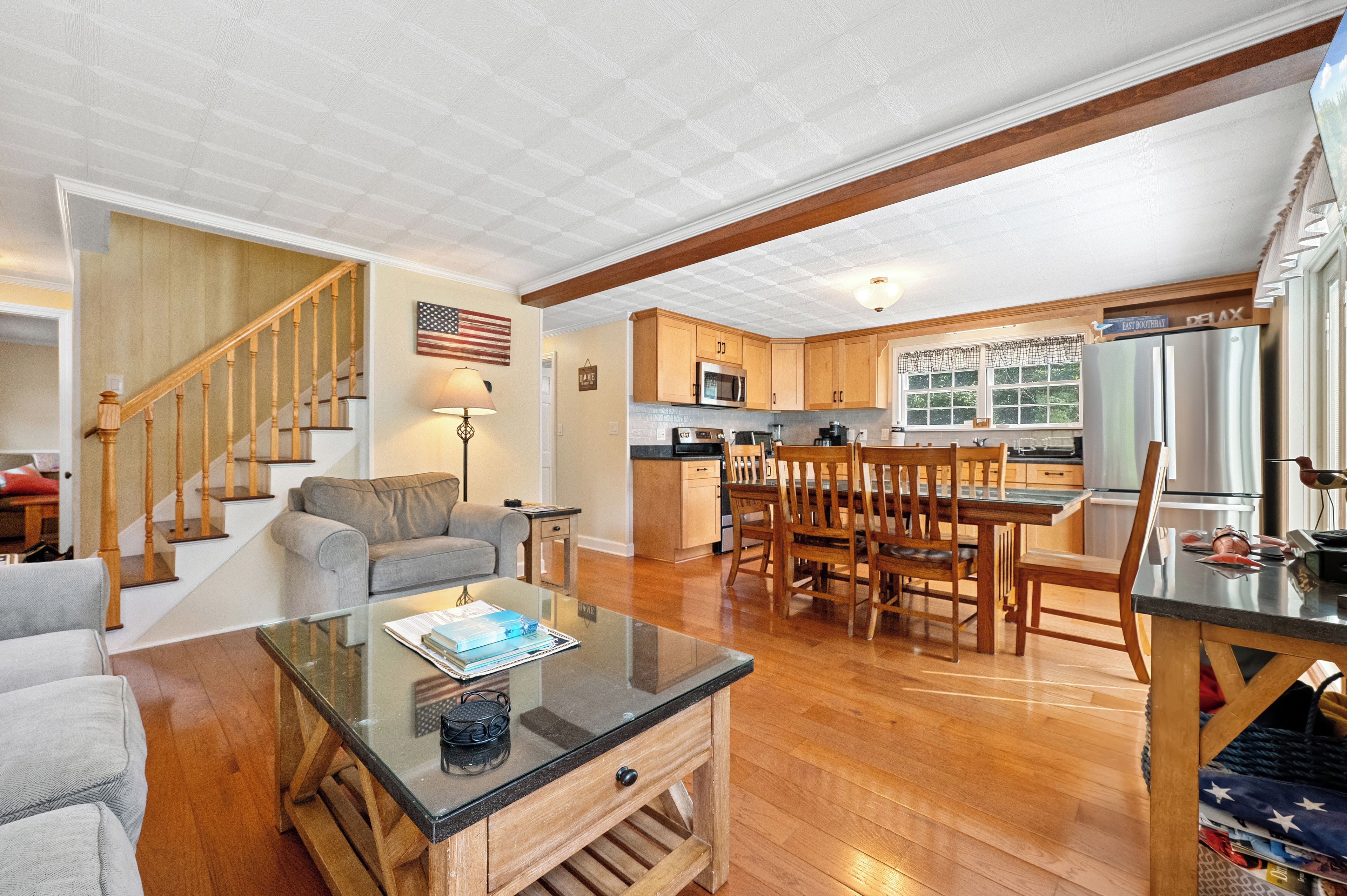 A spacious living area with a glass coffee table, wooden dining table, and a kitchen with wooden cabinets and a stainless steel refrigerator.