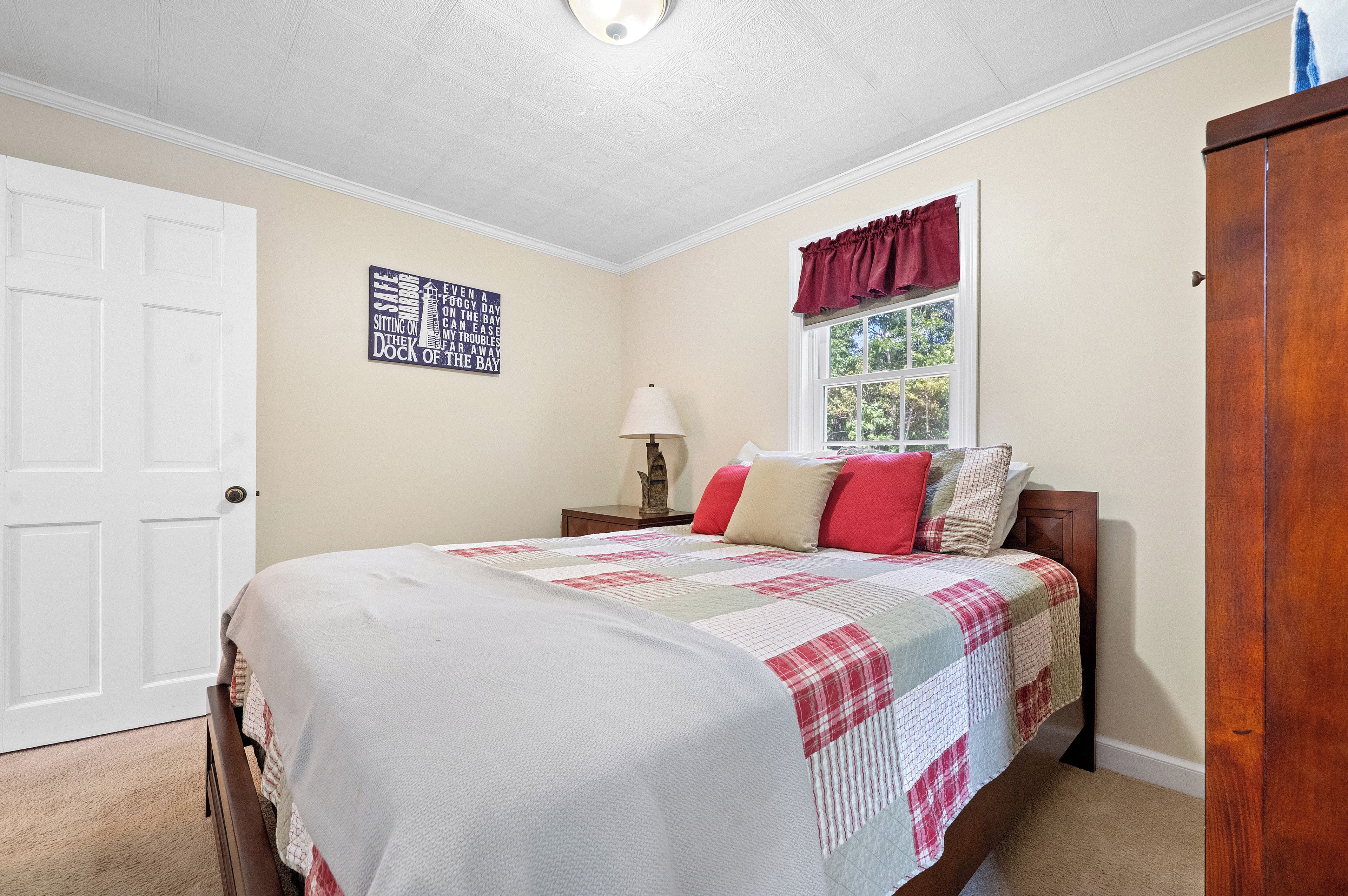 A bedroom with a bed, a window with curtains, a nightstand, a lamp, and a wall sign.