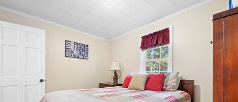A bedroom with a bed, a window with curtains, a nightstand, a lamp, and a wall sign.