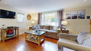A living room with a sofa, coffee table, TV, and artwork on the wall. - Boothbay Harbor Home with Water Views | Near Dining & Whale Watching (Boothbay Harbor)