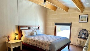 Ein Schlafzimmer mit einem hölzernen Bett, einem Nachttisch mit Lampe, einem Fenster mit Blick und einem Ventilator.