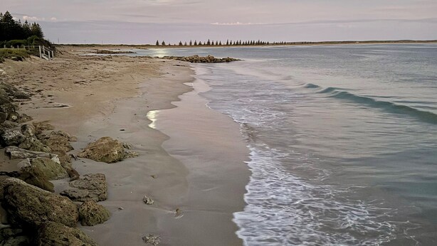Ein Strand mit felsigem Ufer, ruhigem Wasser und einer entfernten Baumgrenze.