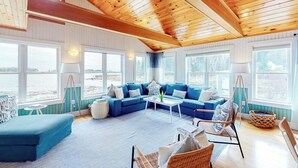 A spacious living area with a blue sofa, a coffee table, and wicker chairs. Large windows offer a view of the outdoors.