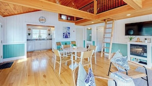 A spacious room with a wooden ceiling, a dining area with chairs, a kitchen with white cabinets, and a TV.