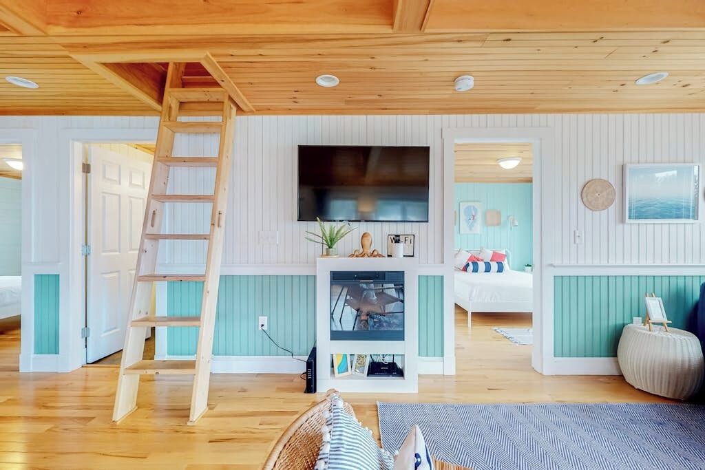Une pièce avec une échelle en bois, un téléviseur à écran plat, un petit téléviseur posé sur un meuble, un lit, un canapé et une horloge fixée au mur.