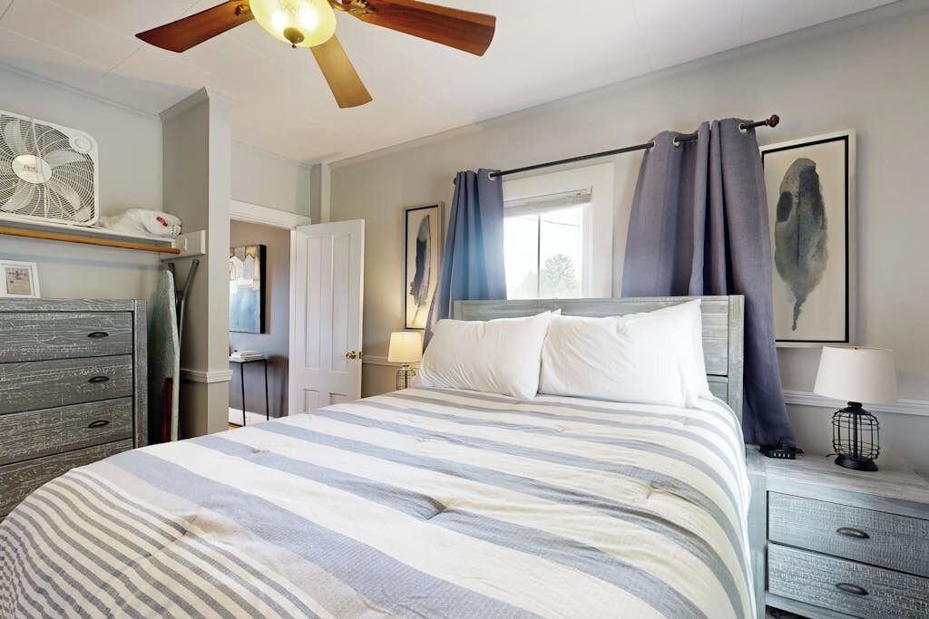 Ein Schlafzimmer mit Bett, Deckenventilator, Kommode, Nachttisch und Fenster mit Vorhängen.