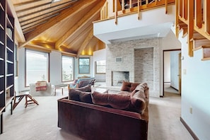 A spacious living room with a high ceiling, a stone fireplace, and a large window. - Private Oceanfront Home -- 2 Fireplaces, 4 Decks & Grill, Dog-friendly (East Boothbay)