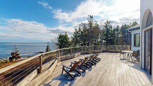 Um deck de madeira com cadeiras e mesas, com vista para o oceano e árvores.