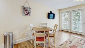 Un comedor con una mesa redonda y cuatro sillas, un televisor de pared, un bote de basura y una puerta con paneles de vidrio.