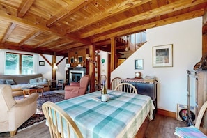 Une pièce chaleureuse avec un plafond en bois, une table à manger recouverte d’une nappe à carreaux, un canapé, une cheminée et un escalier menant à un étage supérieur.