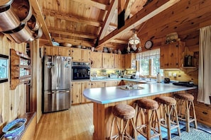 A wooden cabin kitchen with a large island, bar stools, a refrigerator, and a stove. - Waterfront Home on Moose River with Firepit, Porch, Pool Table, Great River View (Rockwood)