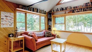A cozy room with a red sofa, a small table, a bookshelf, and large windows offering a view of trees. - Dog-Friendly Lakefront Home w/ Deck, Firepit, Water Views & Games (Rockwood)