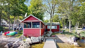 A red cabin with a dock leading to a clear stream, surrounded by trees and other cabins.