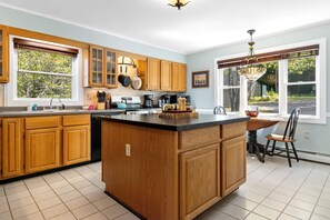 A kitchen with wooden cabinets, a large island, a dining area with a chair, and a window with a view of trees. - Waterfront Oasis - Charming home with stunning bay views, sunroom, gazebo, deck (Trenton)