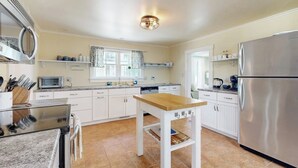 Una cocina con armarios blancos, un refrigerador de acero inoxidable, una mesa de madera y una ventana con cortinas.