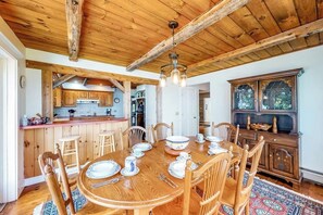 Une table de salle Ă manger en bois, dressĂ©e avec des assiettes, des tasses et des couverts, entourĂ©e de chaises, dans une piĂšce aux poutres apparentes et dotĂ©e dâune grande fenĂȘtre.