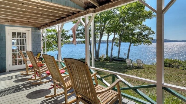 A covered deck with wooden chairs, a glass door, and a view of a lake. - Spacious Waterfront Estate & Cabins W/ Porch, Fireplace, Kayaks & Free WiFi (Brunswick)