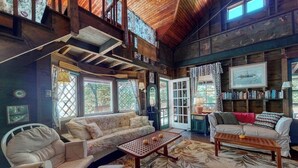 A cozy cabin interior with a high ceiling, wooden beams, a large window, and a bookshelf. - Spacious Waterfront Estate & Cabins W/ Porch, Fireplace, Kayaks & Free WiFi (Brunswick)