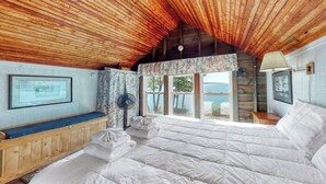 A bedroom with a wooden ceiling, a large bed, a bench, a fan, and a window with a view. - Spacious Waterfront Estate & Cabins W/ Porch, Fireplace, Kayaks & Free WiFi (Brunswick)
