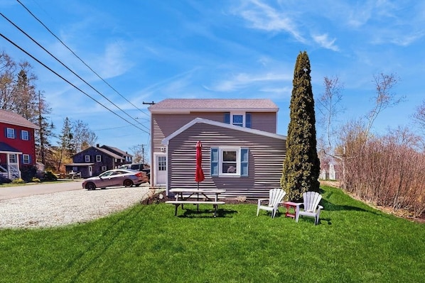 A two-story house with a red car parked in the driveway, a picnic table, and two Adirondack chairs in the front yard.
