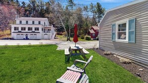 En gräsyta med ett picknickbord och två Adirondack-stolar, ett vitt hus med ett garage och en röd husvagn.